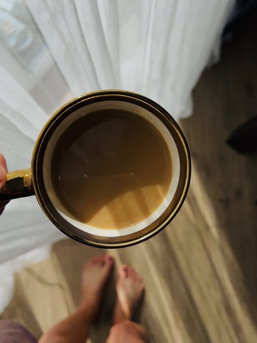 A top-down view of a hand holding a nearly empty coffee mug with brown coffee residue, standing barefoot on a wooden floor in front of white sheer curtains.