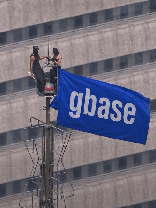 Two people wearing black masks and clothing are perched on a tall pole or tower, securing a large blue flag that reads "gbase" in white lowercase letters, set against the grid-like facade of a skyscraper.
