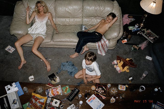 A photograph dated "MAY 15 '86" in red text, depicting a messy living room with a woman in a white slip on a couch, a shirtless boy in jeans, another young person on the floor, and a coffee table cluttered with magazines, remotes, and trash.