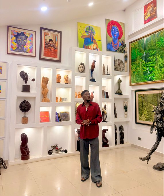 A man in a red shirt and grey pants stands in an art gallery surrounded by colorful paintings on the walls and various wooden sculptures displayed in illuminated alcoves.