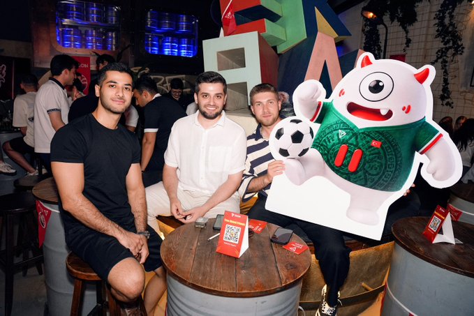 Three young men pose with a soccer ball next to a large white cat mascot wearing a green jersey at what appears to be a FIFA event venue.