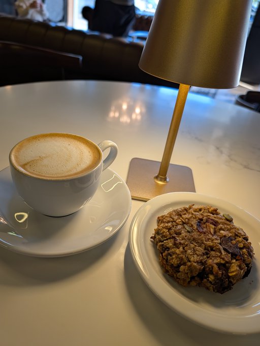 Cozy café scene with a cappuccino in a white cup on a saucer and a homemade-looking oatmeal raisin cookie on a plate, set on a marble table beside a small gold table lamp.