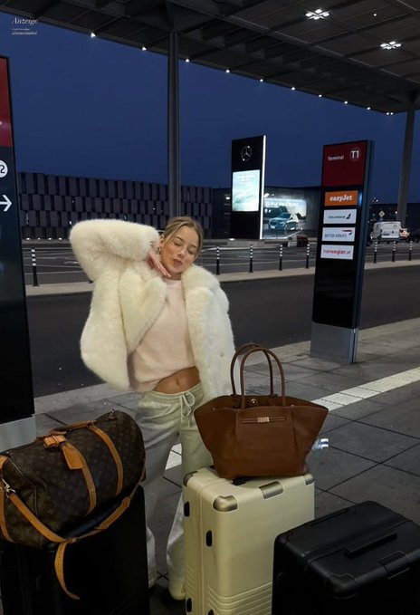 A woman in a fluffy white jacket poses with luggage, including a Louis Vuitton duffel bag, outside an airport terminal at dusk with signs for Terminal 1 and easyJet visible.
