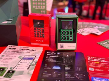 A promotional display showing a green KeyPal TokenPocket hardware device on a red surface alongside product brochures, with text mentioning 'USDD TokenPocket' and '$1,000,000 REWARD'.
