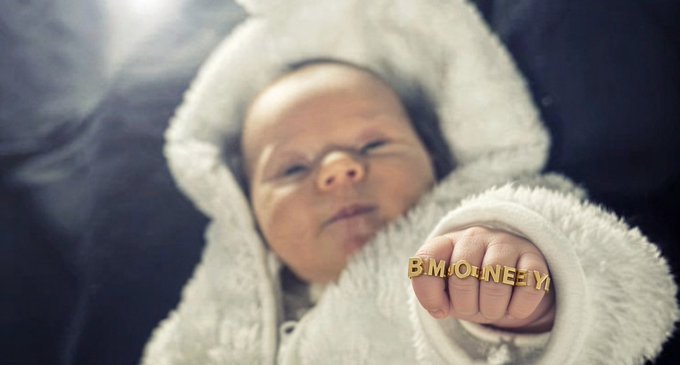 A close-up photograph of a sleeping newborn baby wearing a white knit hooded outfit, with a gold 'BMONEY' bracelet displayed on its tiny hand.