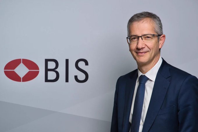 A man in a navy blue suit and tie stands smiling beside the red "BIS" logo (Bank for International Settlements), visually representing the institution warning about stablecoin risks and advocating for tokenized deposits as a payment alternative.