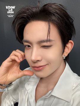 A young man winks playfully at the camera while resting his hand near his face, wearing a white collared shirt and small earrings. The image features "KCON LA 2025" text in the upper left corner, and it accompanies a post promoting KCON LA 2026, with the caption teasing a performer named EVAN for the upcoming event.