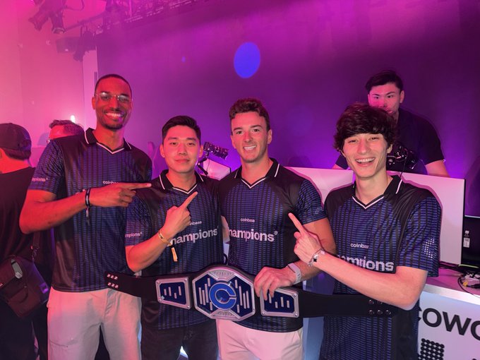 Four teammates wearing matching blue "Champions" jerseys pose together at an event, with two holding up a large championship belt that reads "CHAMPION" across the center. The image aligns with the caption celebrating Team America's victory, showing the winning squad proudly displaying their championship title in a vibrant, pink-lit venue.