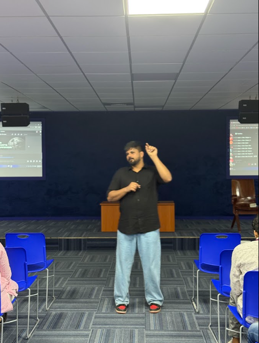 A man in a black shirt and jeans stands on a stage holding a microphone and gesturing during a presentation in a room with blue chairs.