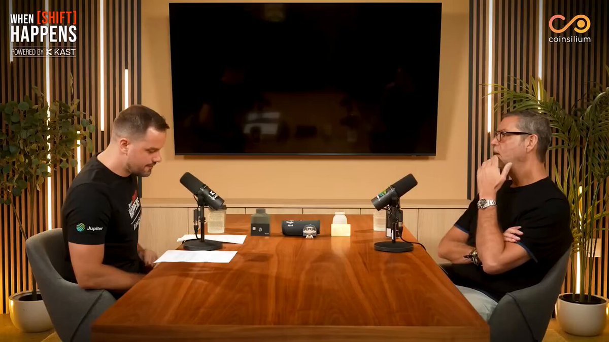 The image features two men sitting at a table, each holding a microphone, suggesting they are hosting a podcast or radio show. The hosts appear to be discussing the concept of digital assets and their value. The background includes a television screen mounted on the wall behind them, which could potentially be used for visual content during their broadcast.
