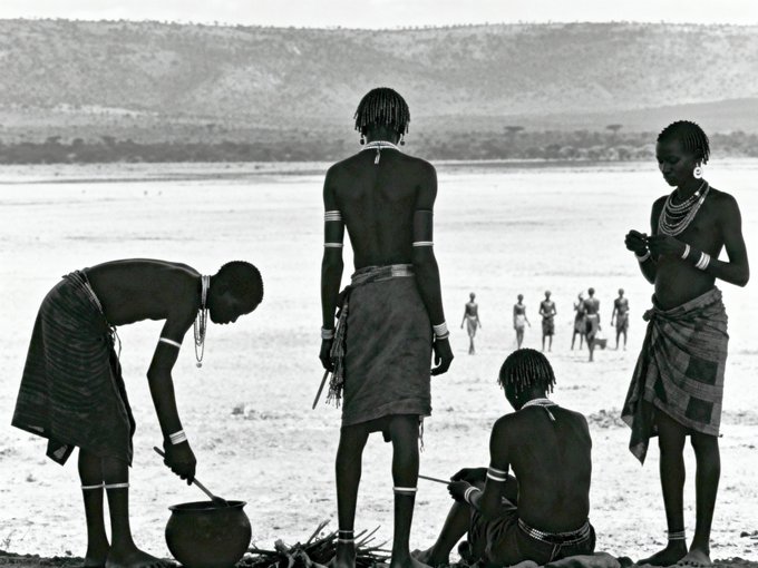 A black-and-white photograph of African people in traditional dress standing and sitting near a body of water, with one person tending a pot over a small fire.