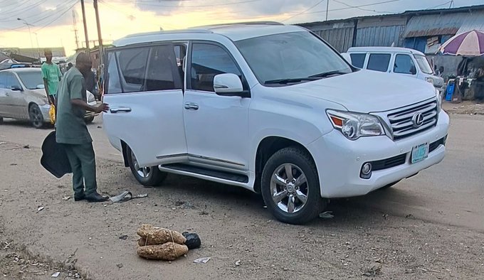 A white Toyota SUV parked on a dirt area with a man in a green shirt standing by the open driver's door, with woven baskets on the ground nearby in what appears to be a West African setting.