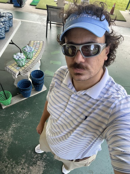 A selfie of a man wearing a blue H.S. Playera visor, sunglasses, and a striped polo shirt, standing at a golf driving range with baskets of golf balls.