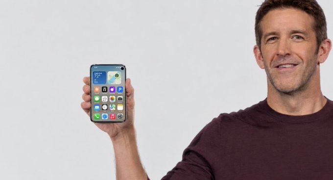 A man with short brown hair, wearing a maroon shirt, smiles while holding up a smartphone displaying its home screen with various app icons. The image ties to "Familiar faces" by showcasing a person presenting a new or recognizable device, suggesting a product introduction or review.