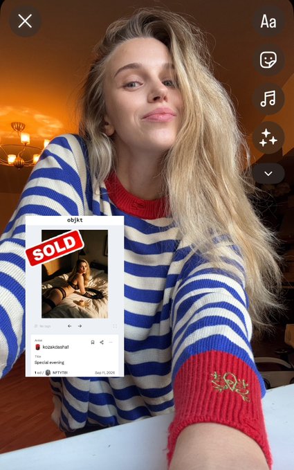 A selfie of a blonde woman in a blue and white striped sweater with red cuffs, posing with a sticker overlay of a 'SOLD' post from 'objkt' by 'kirstenrafael'.