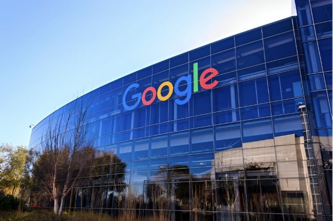 A photograph of a modern glass building featuring the colorful Google logo on its facade, reflecting the blue sky and surrounding trees.