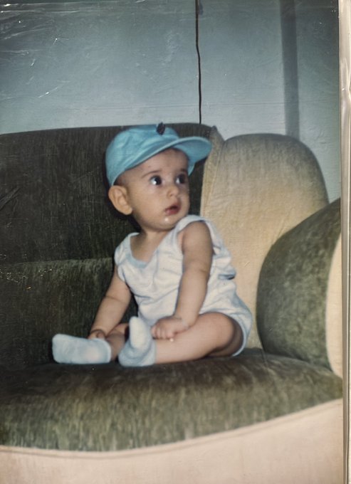 A vintage photograph of a baby wearing a blue cap and light-colored outfit sitting on a green couch.