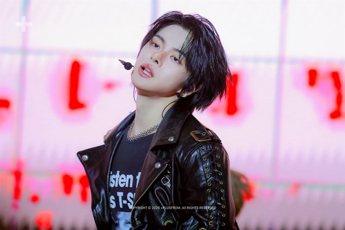 A performer with shoulder-length dark hair, wearing a black leather jacket over a graphic tee, is captured mid-performance on stage with a pink-lit backdrop featuring large letters and the date "260725." The caption "#LEO #리오" identifies this as a stage moment featuring the artist LEO.