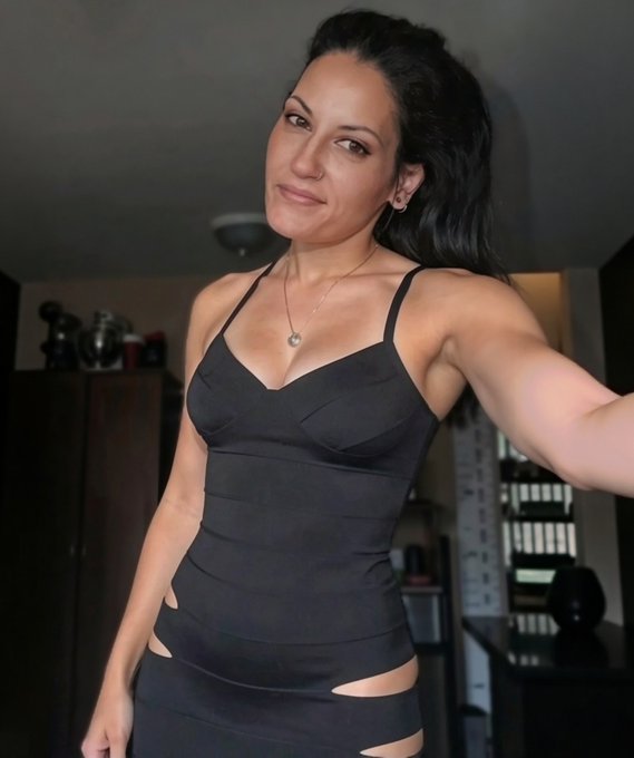 A woman with dark hair pulled back, wearing a black sleeveless dress with cut-out details, a heart pendant necklace, and smiling at the camera in an indoor setting.
