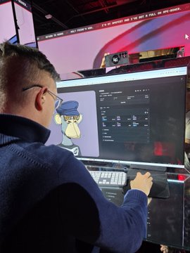A man wearing glasses viewed from behind, working at a dual-monitor computer setup with a cartoon character wallpaper visible on one screen, in what appears to be a studio or tech environment.