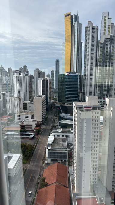 A cityscape view from a high-rise window showing modern skyscrapers, including a distinctive gold-colored building, alongside a street with cars and lower buildings below.