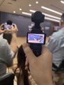 A hand wearing a black glove holds up a smartphone displaying a small screen, capturing a moment at a Binance event where CZ discusses blockchain education in universities.