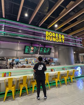 A customer in a Mickey Mouse shirt stands at the counter of a "Bored & Hungry" restaurant, where vibrant green neon lights illuminate the dining area, matching the post's "seeing green" theme.