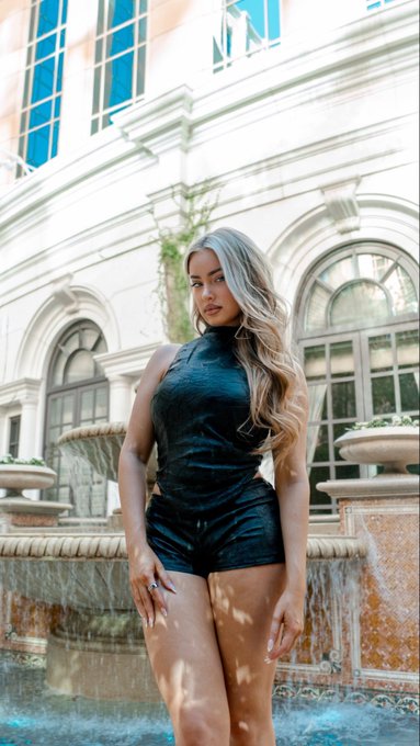 A woman with long, wavy blonde hair poses in a sleek black halter romper in front of an elegant white building with arched windows and a tiered stone fountain, embodying a glamorous "GM gang" morning vibe as part of her 189th day of a 365-day photo project.