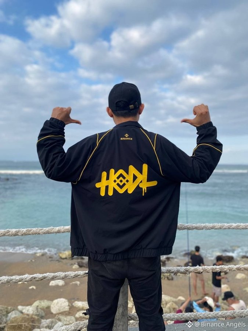 A person wearing a black Binance jacket with 'HOH' in yellow graffiti-style lettering on the back, posing with thumbs down at a beach, credited to '@Binance_Angels'.