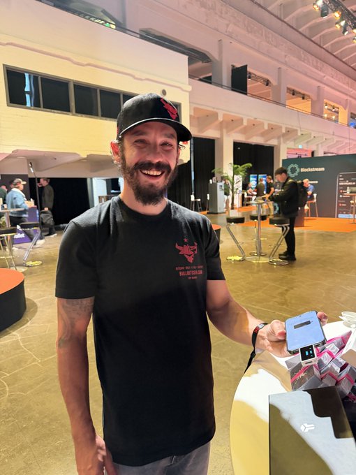 A smiling bearded man wearing a black cap with a red Bitcoin logo and a black "Bitcoin Bottom" t-shirt stands at what appears to be a Bitcoin conference venue, holding a BitBox hardware wallet in one hand and a phone displaying the Bull Bitcoin mobile wallet app in the other, demonstrating the cross-device pairing capability mentioned in the post.