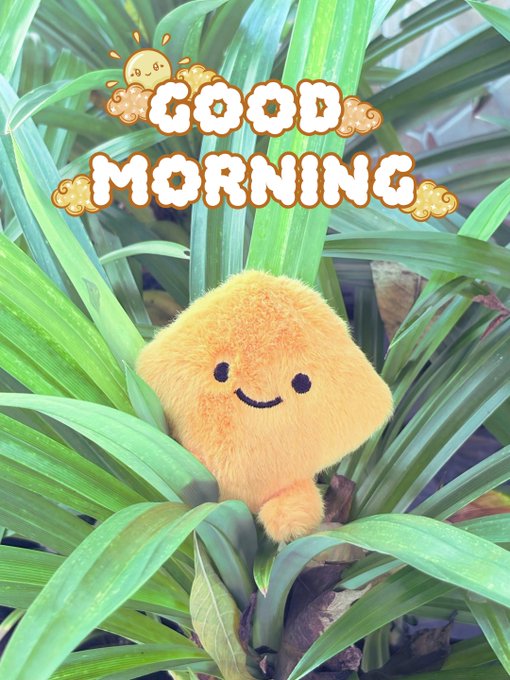 A cheerful 'GOOD MORNING' greeting with pixel-art sun graphics over a small orange smiley plush toy nestled among green plant leaves.
