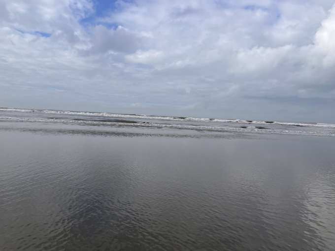 A serene beach scene showing gentle waves rolling onto wet sand under an expansive sky filled with scattered clouds.