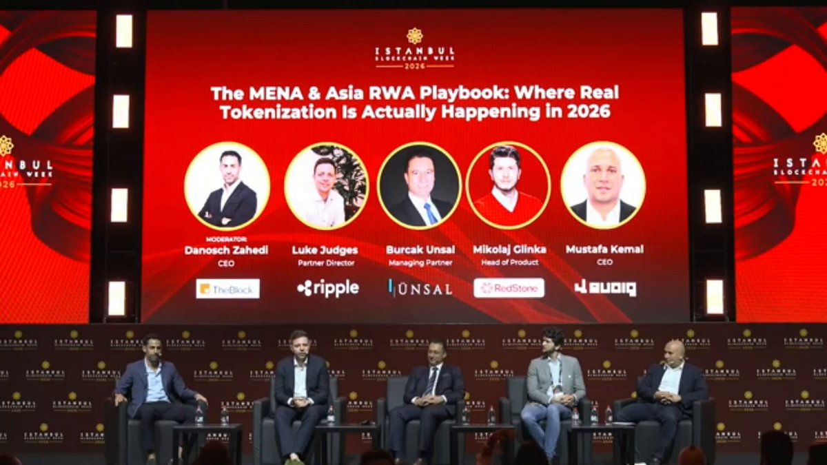 The image shows a group of five men sitting around a table, with one man at the head of the table holding a microphone. They are all dressed professionally and appear to be participating in an event or panel discussion. The backdrop features a large screen displaying text related to Ripple X Ledger and RWA Playbook, indicating that they might be discussing the topic of tokenization and its impact on the financial industry.