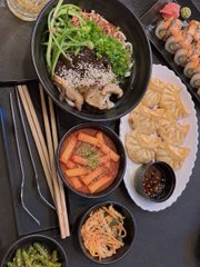 An overhead view of an Asian meal featuring bowls of noodles with mushrooms and greens, a bowl of spicy red soup, dumplings, sushi, pickled vegetables, and chopsticks.
