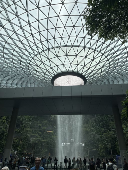 The interior of Jewel Changi Airport features a massive indoor waterfall, with the circular oculus of the glass and steel dome roof visible above.