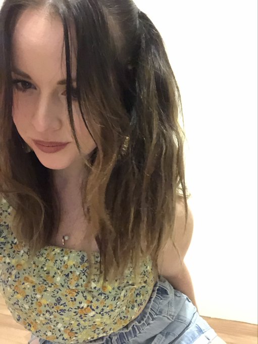 A young woman with long wavy brown hair wearing a yellow floral sleeveless top and light blue denim shorts, posing indoors against a white wall.