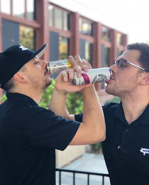 Two men in black polo shirts clink and drink from White Claw Hard Seltzer cans while standing on an outdoor balcony.