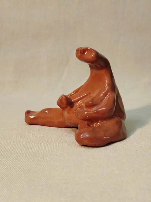 A glossy, reddish-brown clay sculpture of a seated figure with abstract, flowing forms rests on a light fabric surface.