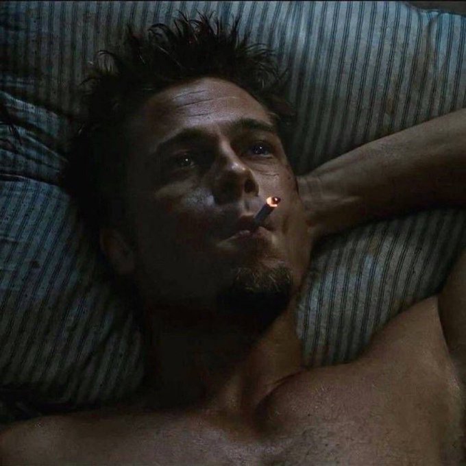 A dimly lit cinematic still of a shirtless man lying on striped bedding, smoking a cigarette with a pensive expression (appears to be Edward Norton in Fight Club).
