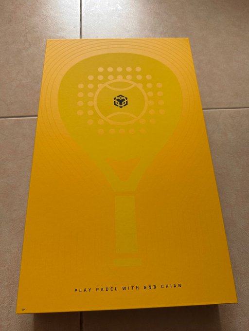 A yellow promotional card or poster on a tiled floor featuring a stylized graphic of a padel racket and the BNB Chain logo, with the text 'PLAY PADEL WITH BNB CHAIN' at the bottom.