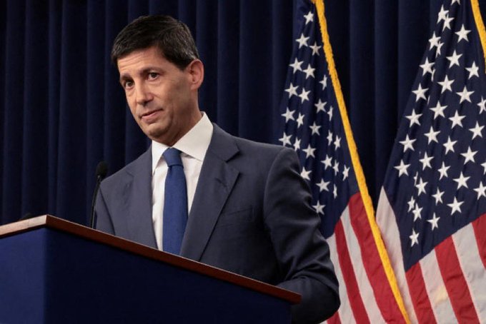 A man in a dark suit and blue tie standing at a podium with microphones, flanked by two American flags.
