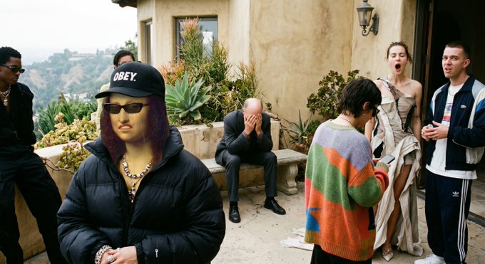 A group of people gathered on a patio/balcony with a hillside view in the background, featuring a person in the foreground wearing a black 'OBEY' cap, dark sunglasses, and a puffy jacket, while others stand around in casual clothing.