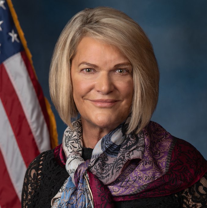An official-style portrait shows a smiling woman with shoulder-length blonde hair, wearing a black lace top and a purple and blue patterned scarf, posed in front of an American flag.

The image likely relates to the Crypto CLARITY Act news, as the woman pictured appears to be Senator Cynthia Lummis (R-WY), a prominent Republican known for her advocacy on cryptocurrency legislation.