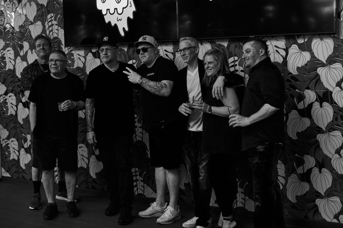 A black-and-white photo of six people posing together indoors against a floral-patterned wall, with one man wearing a New York Yankees cap.