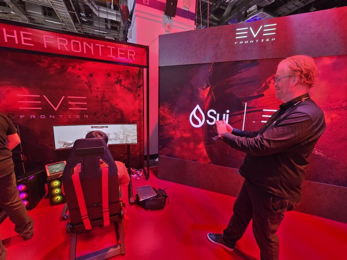A man in glasses stands in a red-lit booth at an EVE Frontier gaming event, gesturing with his hands while a seated gamer plays on a racing seat setup in front of an 'EVE Frontier' screen.