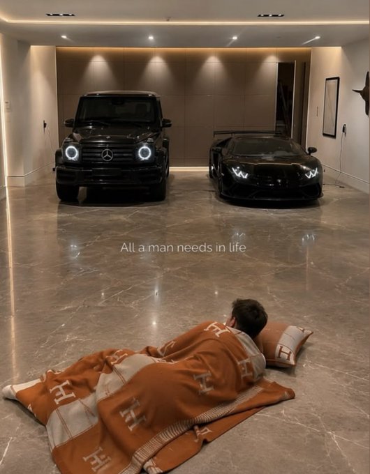 A young boy sleeps peacefully on an Hermès blanket and pillow in a luxurious garage, gazing at a black Mercedes G-Wagon and a black Lamborghini Huracán, embodying the caption's message that "all a man needs in life" is luxury cars and comfort.
