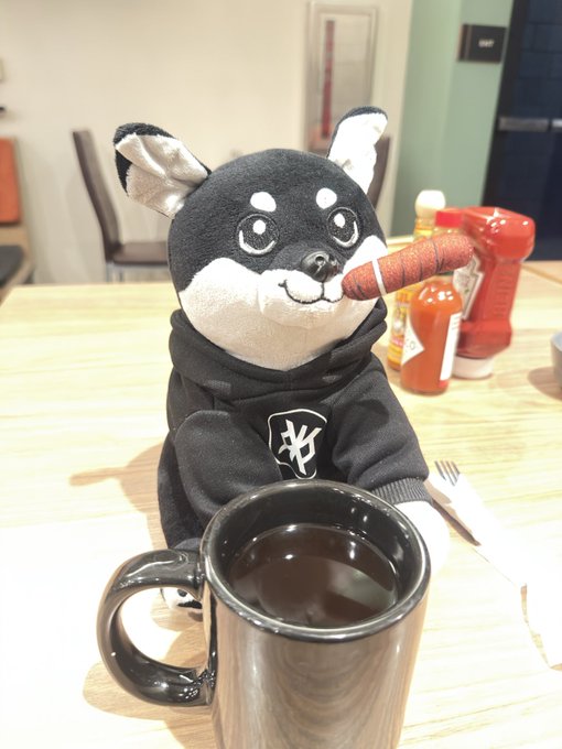 A black and white shiba inu plush toy wearing a hoodie holds a fake carrot in its mouth while sitting at a wooden table next to a black mug of coffee.