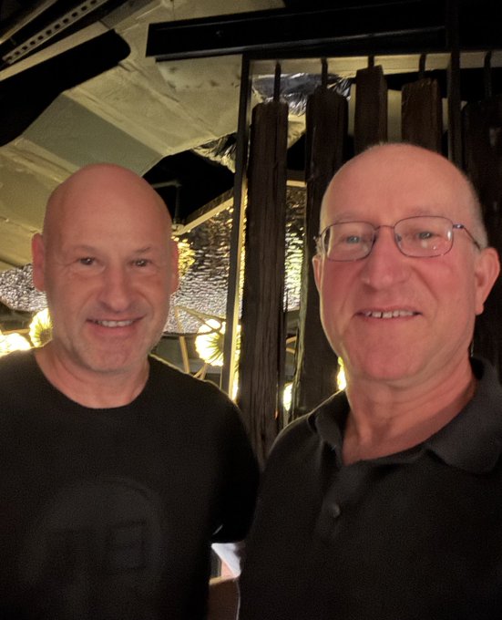 Two smiling, bald men posing together indoors; the man on the right wears glasses and a gray shirt, while the man on the left wears a black t-shirt, with decorative lighting visible in the background.