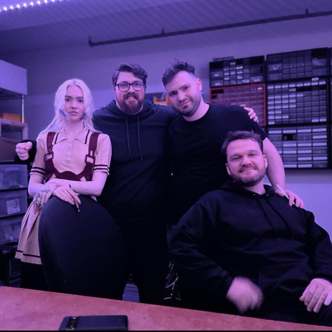 Four people pose together in a room with purple lighting and storage shelving in the background, suggesting a behind-the-scenes setting for the story mentioned in the caption involving collaborators @Grimezsz and @shawmakesmagic.