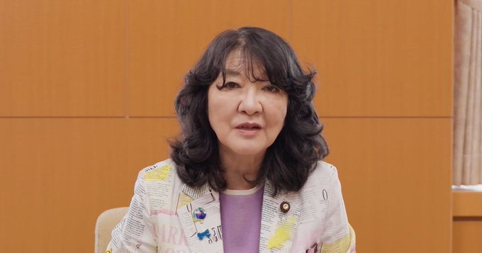A woman with shoulder-length dark wavy hair, wearing a patterned jacket over a purple top, speaks in what appears to be a formal interview or press setting with a wooden panel background. Based on the accompanying Japanese text about cryptocurrency ETF regulations and tax reform, the image likely depicts Financial Services Minister Satsuki Katayama discussing Japan's plans to permit crypto asset ETFs.
